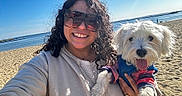 Frankie is registered to the contest to win money with this photo: person, dog, beach, sand, water, ocean, sunny, smile, sunglasses, curly_hair, pet, outdoor, casual_clothing, happy, portrait, holding, blue_sky, jacket, daytime, leisure