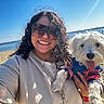person, dog, beach, sand, water, ocean, sunny, smile, sunglasses, curly_hair, pet, outdoor, casual_clothing, happy, portrait, holding, blue_sky, jacket, daytime, leisure