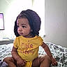 Azori is registered to the contest to win money with this photo: toddler, child, baby, bed, blanket, yellow_shirt, bonnet, indoor, window, natural_light, patterned_bedding, cute, sitting, person, portrait, clothing, young_child, curious, casual, home