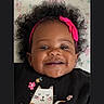baby, smiling, headband, pink_bow, curly_hair, black_shirt, cat_illustration, flowers, cute, portrait, child, happy, earrings, indoor, blanket, closeup, face, infant, apparel, cozy
