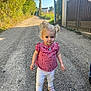 toddler, child, girl, blonde_hair, pigtails, red_checkered_top, white_pants, pink_shoes, gravel_path, outdoor, sunny, greenery, fence, gate, walking, person, daylight, nature, portrait, casual_clothing