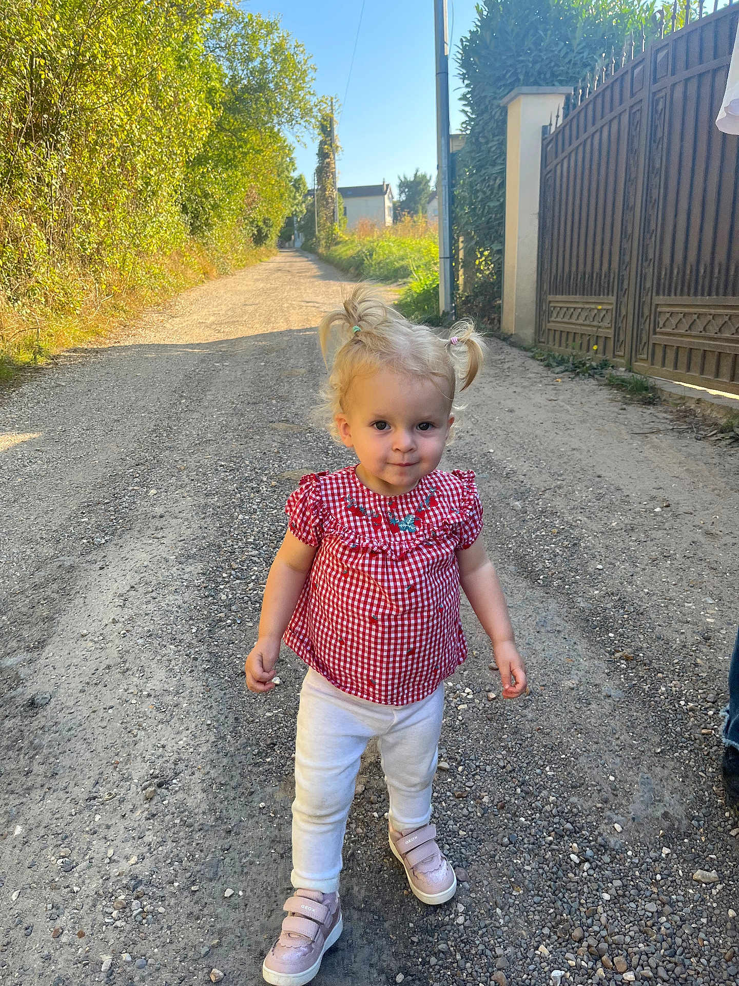 Léna a rejoint le concours — aidez-le/la à gagner de superbes lots ! toddler, child, girl, blonde_hair, pigtails, red_checkered_top, white_pants, pink_shoes, gravel_path, outdoor, sunny, greenery, fence, gate, walking, person, daylight, nature, portrait, casual_clothing