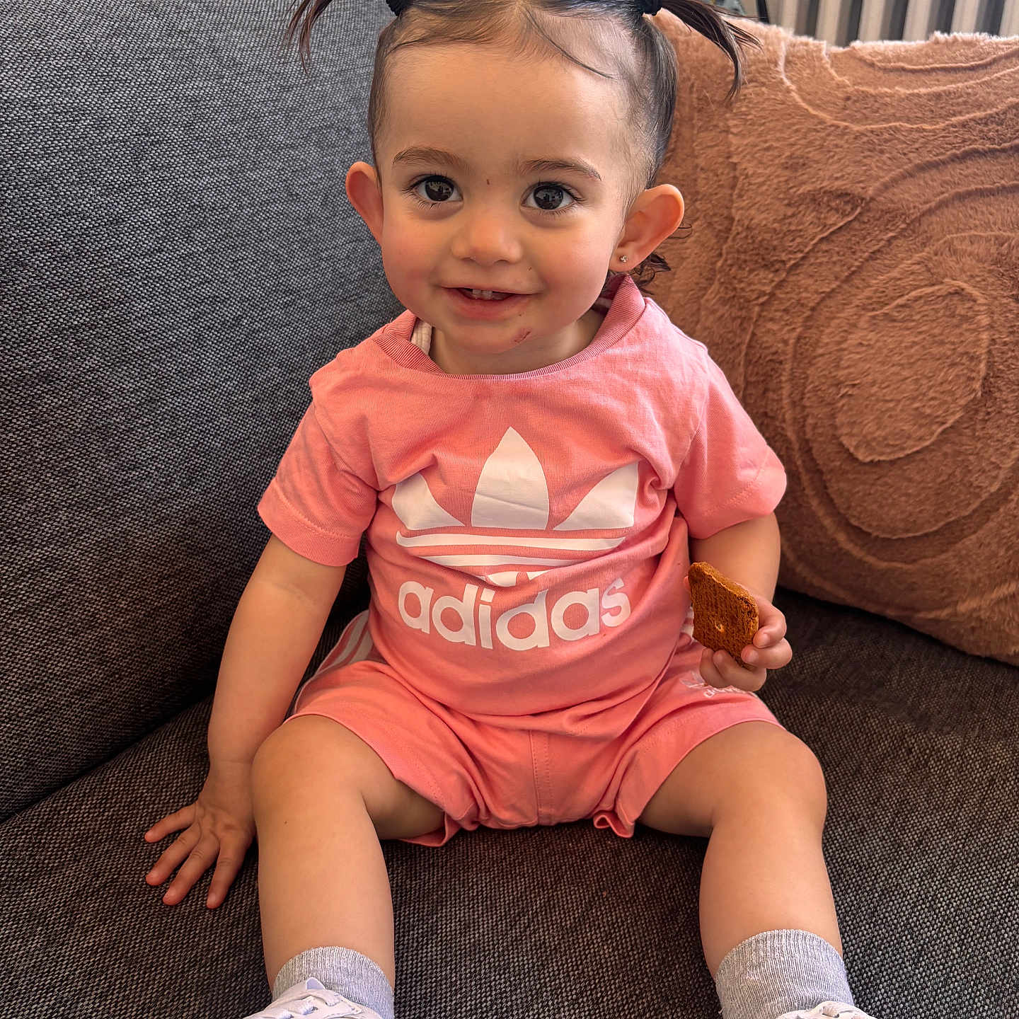 Inaya a rejoint le concours — aidez-le/la à gagner de superbes lots ! toddler, child, pigtails, pink_clothing, adidas, sneakers, white_shoes, couch, gray_couch, brown_pillow, biscuit, smiling, indoor, radiator, happy, seated, cute, young_child, casual, portrait