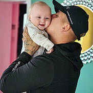 Remi is registered to the contest to win money with this photo: baby, man, kiss, smile, tattoo, cap, under_armour, onesie, indoor, wall, doorway, colorful_background, affection, family, love, holding, portrait, happy, person, head