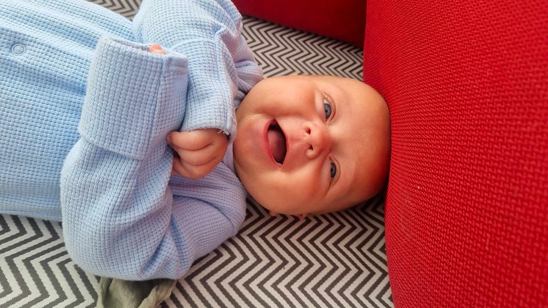 Remi joined the competition — help win amazing prizes! baby, infant, smiling, blue_clothing, red_cushion, patterned_blanket, happy, portrait, closeup, face, child, indoor, cozy, cute, human, laying_down, head, skin, expression, warm