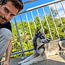man, dog, husky, pet, outdoor, balcony, railing, sunlight, sky, trees, greenery, casual_clothing, beard, smile, portrait, animal, companion, relaxation, daytime, nature
