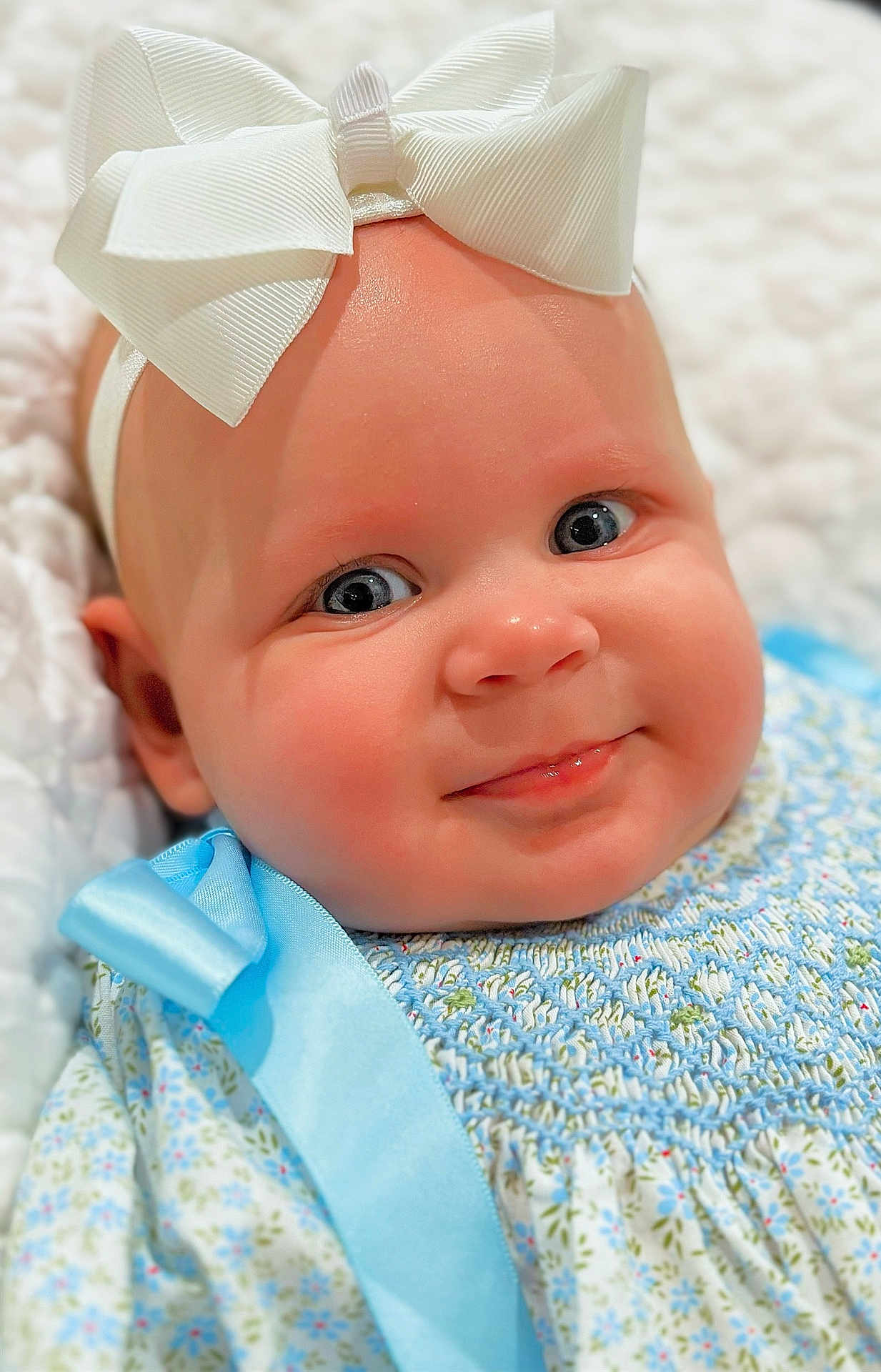 Lynnon is registered to the contest to win money with this photo: baby, infant, face, blue_eyes, bow, headband, smile, dress, blue_ribbon, floral_pattern, soft_blanket, close_up, cute, child, skin, cheeks, portrait, baby_clothing, happy, indoors