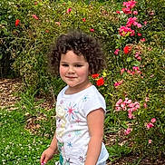 Mila participe au concours pour gagner de l'argent avec cette photo : child, curly_hair, smiling, white_tshirt, flower_garden, pink_flowers, red_flowers, greenery, outdoor, nature, plants, grass, happy, cute, portrait, person, young, summer, daylight, garden_path