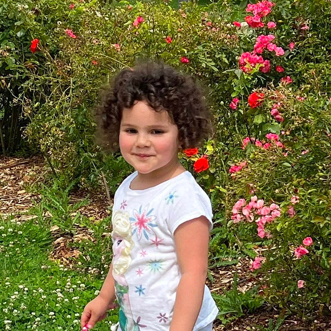 Mila participe au concours pour gagner de l'argent avec cette photo : child, curly_hair, smiling, white_tshirt, flower_garden, pink_flowers, red_flowers, greenery, outdoor, nature, plants, grass, happy, cute, portrait, person, young, summer, daylight, garden_path