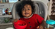 Hector is registered to the contest to win money with this photo: baby, child, toddler, high_chair, smiling, curly_hair, red_clothing, bowl, fruit, strawberries, sippy_cup, kitchen, indoor, home, window, door, table, food, portrait, happy