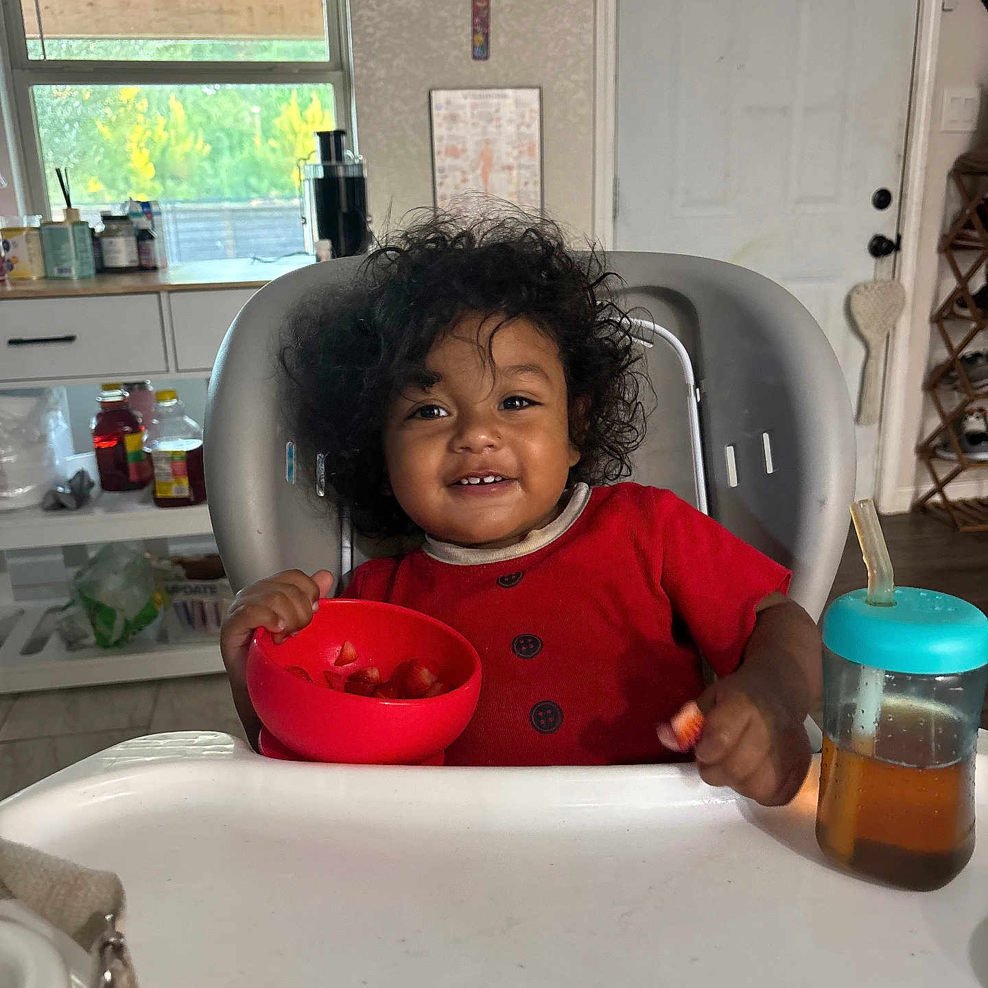 Hector is registered to the contest to win money with this photo: baby, child, toddler, high_chair, smiling, curly_hair, red_clothing, bowl, fruit, strawberries, sippy_cup, kitchen, indoor, home, window, door, table, food, portrait, happy