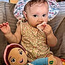 baby, child, pacifier, hat, floral_pattern, plush_doll, indoor, couch, clothing, person, cute, infant, smiling, toy, seated, colorful, face, hand, happy, portrait