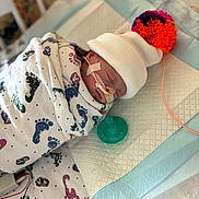 Adalynn is registered to the contest to win money with this photo: newborn, baby, hospital, crib, blanket, footprint_pattern, hat, pom_pom, pacifier, medical_tubes, sleeping, infant, swaddled, cute, healthcare, bed, soft_lighting, closeup, person, resting