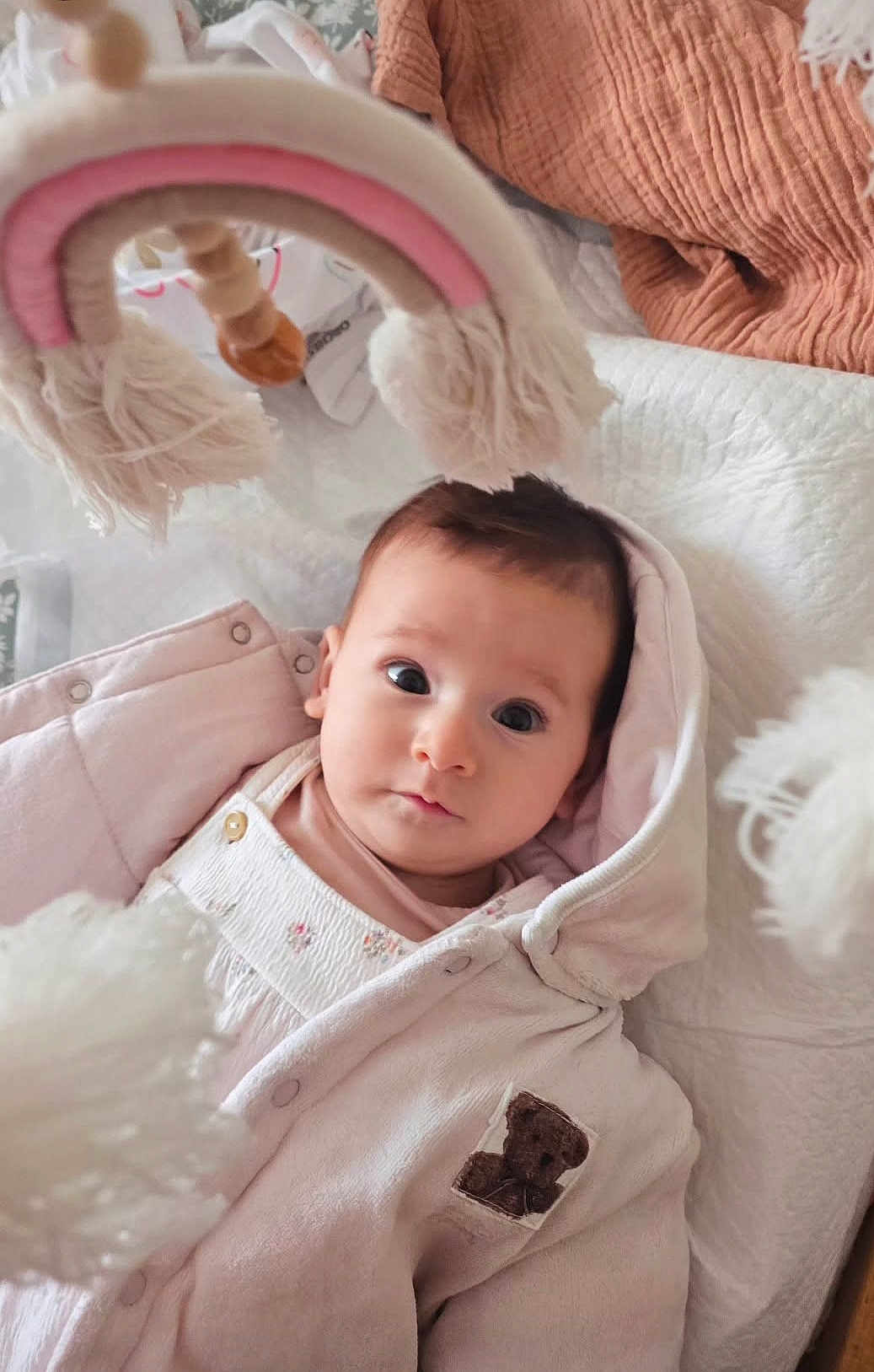 June participe au concours pour gagner de l'argent avec cette photo : baby, infant, child, pink_jacket, hooded_clothing, teddy_bear_patch, toy, rainbow_toy, soft_toy, quilt, blanket, bed, indoor, portrait, face, eyes, cute, cozy, warm, childrens_clothing