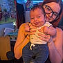 baby, child, woman, glasses, indoor, holding, smiling, tongue_out, jeans, tank_top, led_lights, room, furniture, person, portrait, happy, casual_clothing, red_hair, infant, cute