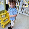 child, toddler, boy, denim_shirt, shorts, black_shoes, yellow_slide, plastic_slide, porch, railing, outdoor, daylight, curly_hair, concrete_floor, greenery, car, fence, playground_equipment, summer, cute
