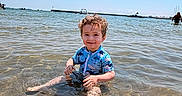 Alex joined the competition — help win amazing prizes! child, toddler, water, beach, sea, sky, sunlight, swimsuit, curly_hair, smile, sitting, shallow_water, sand, outdoor, summer, happy, person, nature, vacation, play