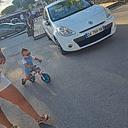 Shelby a rejoint le concours — aidez-le/la à gagner de superbes lots ! toddler, child, bike, balance_bike, sunglasses, white_car, adult, legs, sandals, pavement, outdoor, street, daylight, trees, shadows, people, fence, buildings, summer, casual