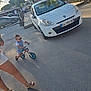 toddler, child, bike, balance_bike, sunglasses, white_car, adult, legs, sandals, pavement, outdoor, street, daylight, trees, shadows, people, fence, buildings, summer, casual