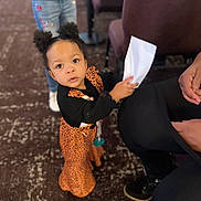 Naomi is registered to the contest to win money with this photo: toddler, child, girl, curly_hair, hair_puffs, orange_dress, black_sleeves, paper, indoor, carpet, chairs, person, looking_up, curious, standing, footwear, jeans, blurred_background, holding_object, expression