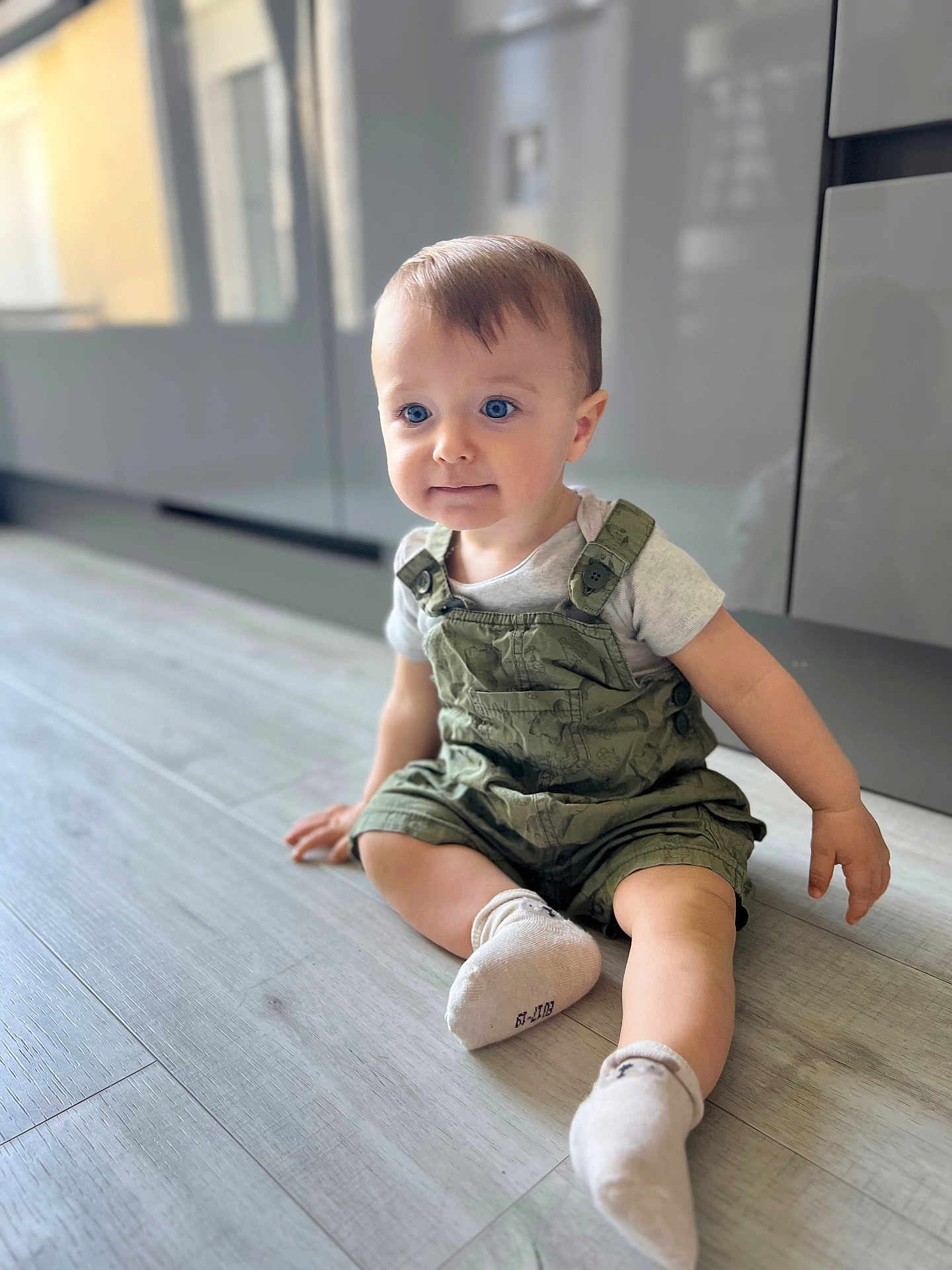 Elio participe au concours pour gagner de l'argent avec cette photo : baby, blue_eyes, cabinet, casual_clothing, child, curious, floor, hair, indoor, infant, kitchen, light, overalls, portrait, sitting, smile, socks, toddler, wooden_floor, young_child