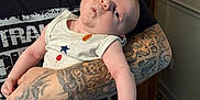 Charles is registered to the contest to win money with this photo: baby, infant, tattoo, arm, person, indoor, clothing, sleeveless_shirt, wooden_furniture, floor, hand, face, expression, curious, holding, skin, casual, adult, portrait, home