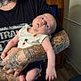 baby, infant, tattoo, arm, person, indoor, clothing, sleeveless_shirt, wooden_furniture, floor, hand, face, expression, curious, holding, skin, casual, adult, portrait, home