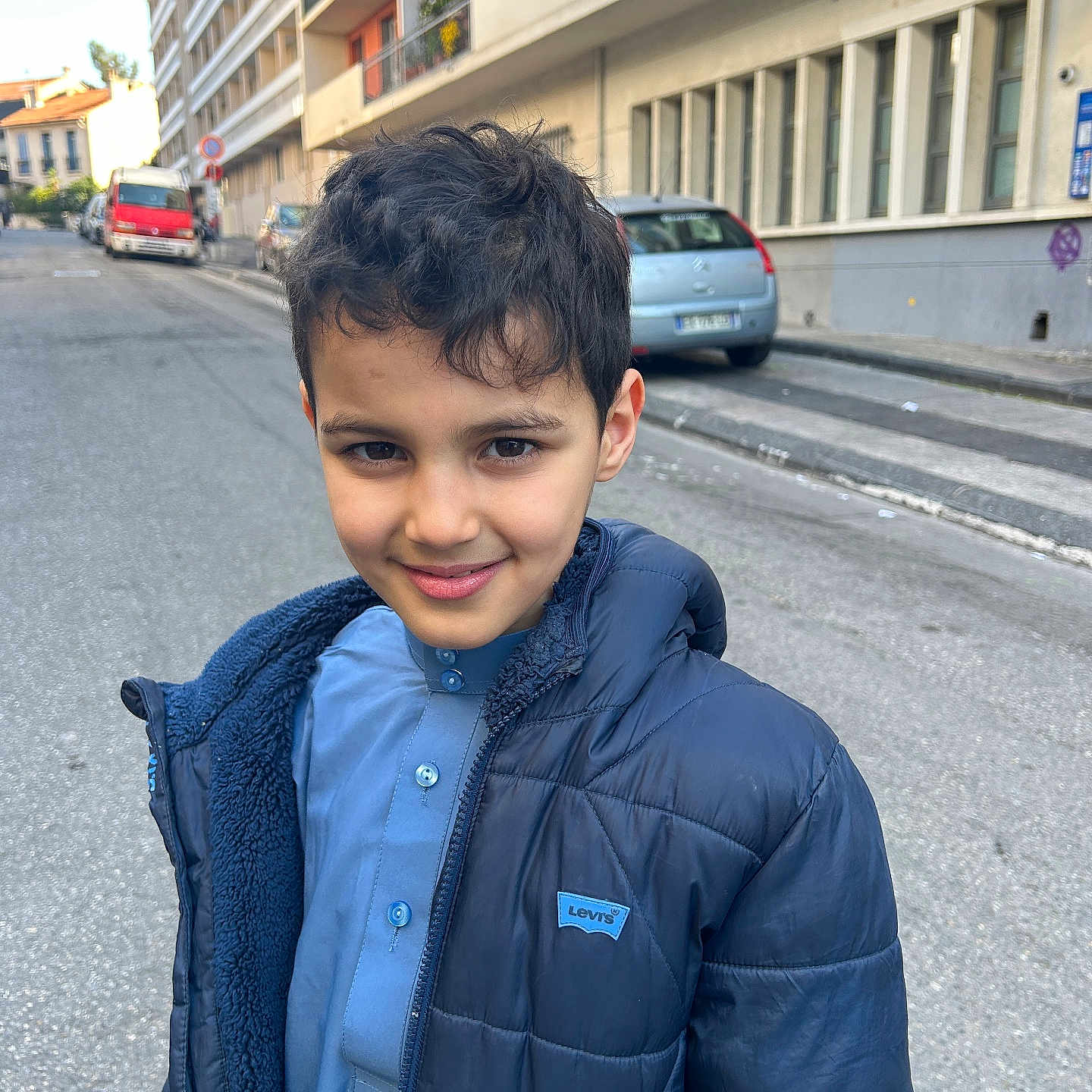 Amir participe au concours pour gagner de l'argent avec cette photo : child, boy, smile, jacket, blue_clothing, urban, street, car, building, sidewalk, daylight, portrait, person, standing, casual, outdoor, young, happy, face, hair
