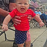 baby, child, infant, smiling, laughing, outdoor, portrait, patriotic, american_flag, red_shirt, star_spangled, shorts, standing, being_held, adult, hands, festival, building, chairs, daylight