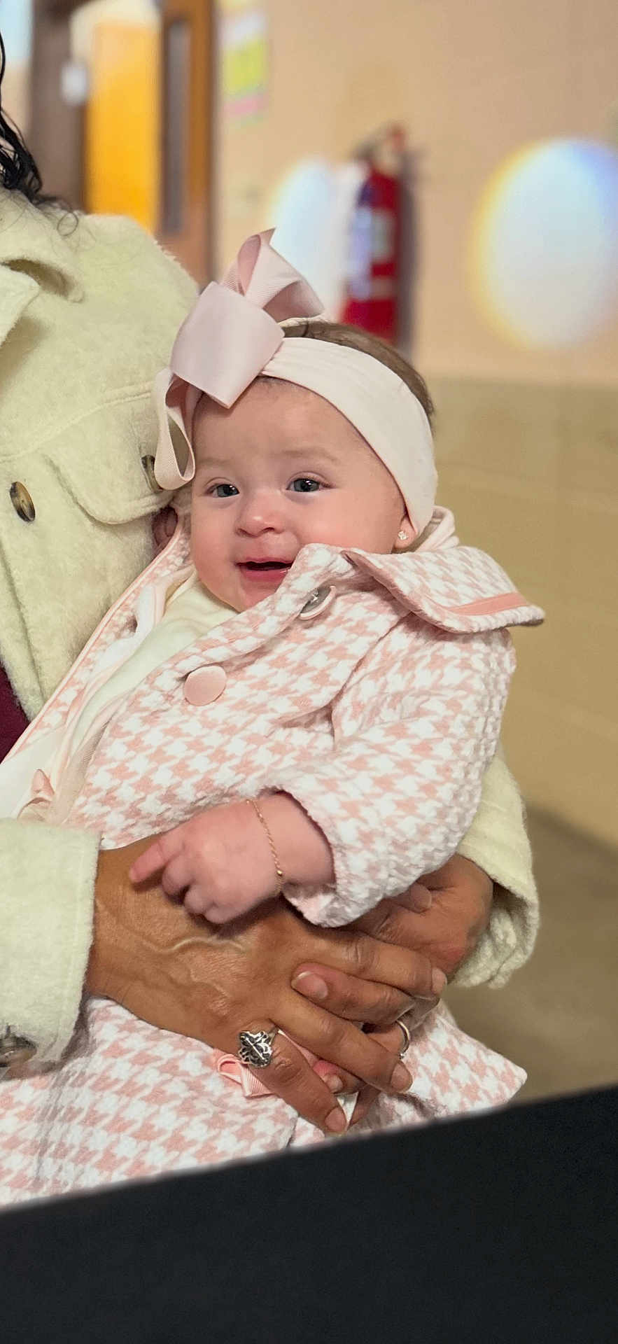 Ariathna is registered to the contest to win money with this photo: adult_hands, baby, blurred_background, bokeh, bow, bracelet, cheeks, coat, earring, fire_extinguisher, hands, headband, holding, houndstooth, indoor, infant, pink, portrait, ring, smile