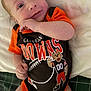 baby, infant, clothing, onesie, sportswear, cleveland_browns, football, cartoon, cute, lying_down, hand, face, eyes, blanket, soft_surface, indoors, child, smile, person, portrait