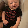 baby, infant, child, smiling, happy, portrait, face, sitting, indoor, cushion, chair, black_shirt, text_on_clothing, red_lettering, bald, eyes, hands, arms, legs, cozy