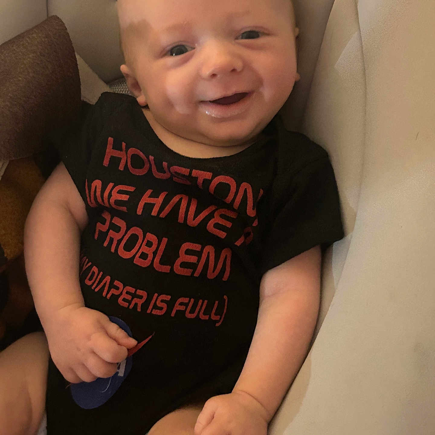 Greysen joined the competition — help win amazing prizes! baby, infant, child, smiling, happy, portrait, face, sitting, indoor, cushion, chair, black_shirt, text_on_clothing, red_lettering, bald, eyes, hands, arms, legs, cozy