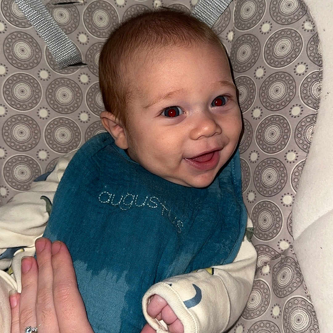 Augustus is registered to the contest to win money with this photo: baby, child, infant, smiling, happy, portrait, bouncer, baby_seat, safety_straps, patterned_fabric, blue_clothing, cream_clothing, hand, adult_hand, indoor, close_up, seated, brown_hair, face, photograph