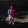 child, girl, basketball, outdoor, court, smiling, sport, playground, shorts, striped_shirt, white_shoes, hair_bows, sunlight, shadow, trees, bench, park, happy, standing, person