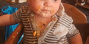 Jasper-Riley is registered to the contest to win money with this photo: baby, casual, child, clothing, curious, cute, expression, face, food, high_chair, indoor, meal, messy, person, seat, small_child, stain, striped_shirt, table, wood
