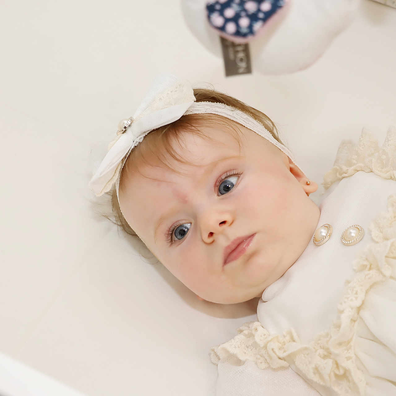 Eva Beatrice a rejoint le concours — aidez-le/la à gagner de superbes lots ! adorable, baby, blue_eyes, bow, child, closeup, curious_expression, cute, earrings, face, frills, headband, indoor, infant, lying_down, person, portrait, skin, soft_lighting, white_clothing