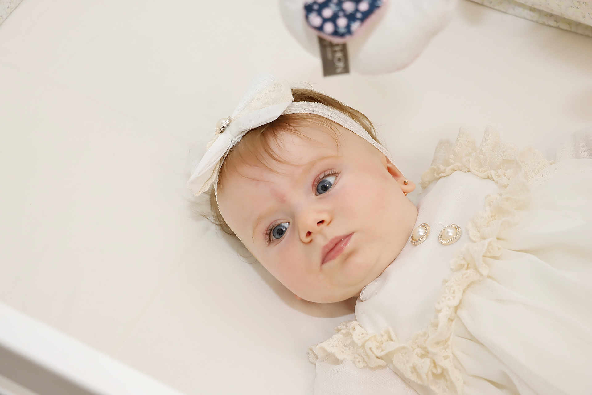 Eva Beatrice a rejoint le concours — aidez-le/la à gagner de superbes lots ! baby, infant, child, portrait, headband, bow, white_clothing, frills, earrings, blue_eyes, soft_lighting, closeup, lying_down, curious_expression, indoor, cute, adorable, face, skin, person