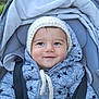 baby, child, smile, face, hat, jacket, puffer_jacket, knitted_hat, stroller, seatbelt, outdoor, cold_weather, cozy, infant, baby_clothing, happy, portrait, headwear, person, cute