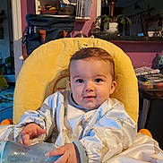 Sacha participe au concours pour gagner de l'argent avec cette photo : baby, child, high_chair, bottle, indoor, yellow_cushion, curious, smiling, clothing, dinosaur_print, face, person, furniture, plant, table, household_items, warm_lighting, background, cozy, portrait