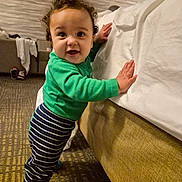 Kyree is registered to the contest to win money with this photo: baby, toddler, child, standing, smiling, green_shirt, striped_pants, curly_hair, bed, bedding, hotel_room, carpet, hand, face, eyes, socks, furniture, indoor, playful, portrait