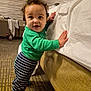 baby, toddler, child, standing, smiling, green_shirt, striped_pants, curly_hair, bed, bedding, hotel_room, carpet, hand, face, eyes, socks, furniture, indoor, playful, portrait