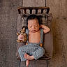newborn, baby, sleeping, teddy_bear, wooden_bed, pillows, knit_overalls, carpet, indoors, cozy, cute, infant, peaceful, resting, relaxed, child, soft_lighting, portrait, tiny_bed, floor