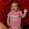 baby, infant, child, smiling, happy, portrait, lying_down, red_blanket, pink_outfit, onesie, sparkly_text, mamas_mini, raised_hand, gummy_smile, indoor, close_up, adorable, fabric, person, family_photo