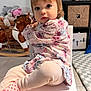 toddler, child, sitting, chair, floral_clothing, pink_pants, polka_dot_socks, indoor, playroom, rocking_horse, stuffed_animals, carpet, curious_expression, young_child, small_chair, portrait, person, floor, home, cute