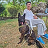 blue_shoes, casual_clothing, chair, daytime, dog, fence, garden, grass, happy_dog, harness, leaves, nature, outdoor, person, plastic_chair, relaxing, rock, smiling, sunlight, tree