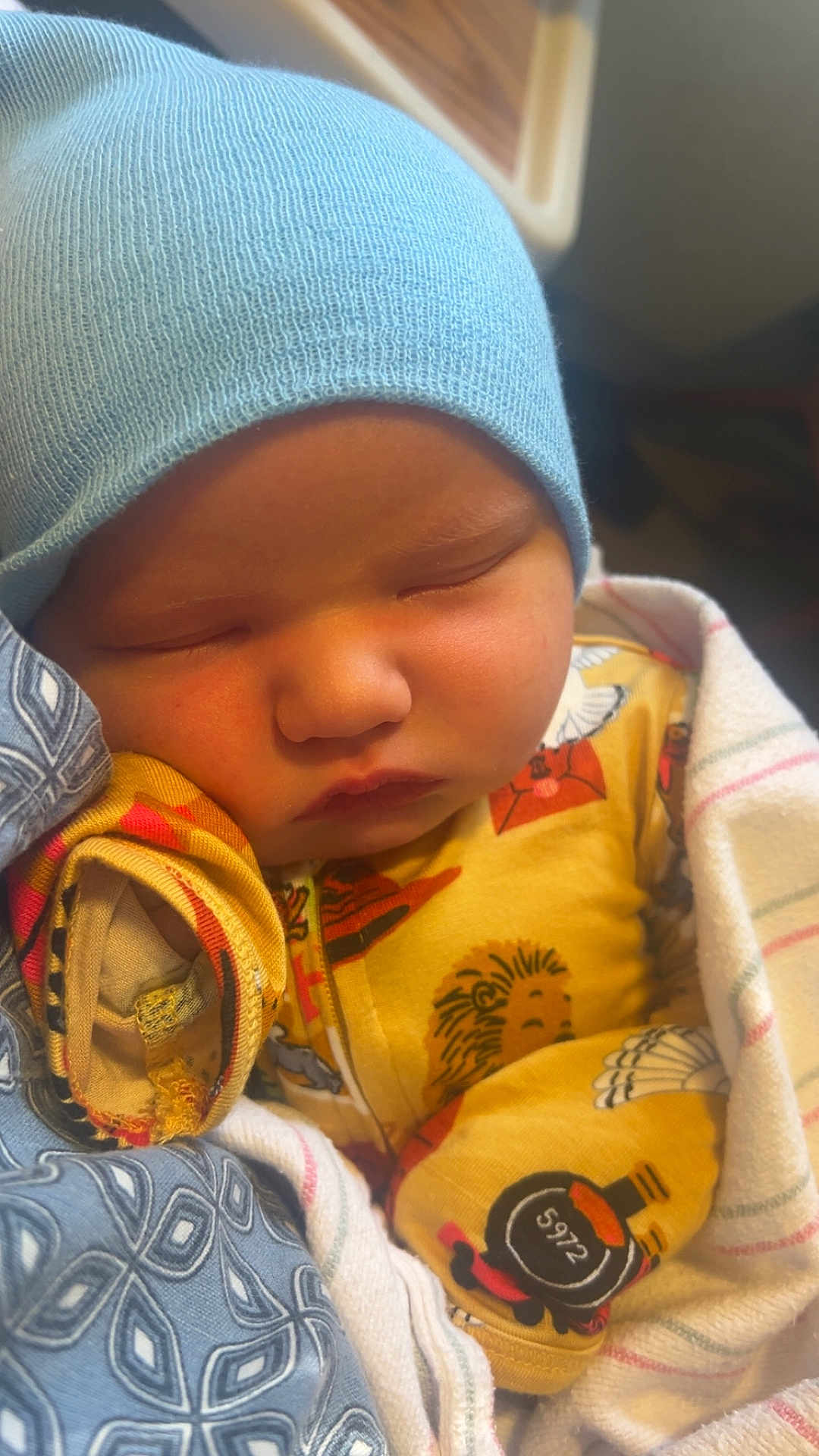Waylon is registered to the contest to win money with this photo: baby, sleeping, blue_hat, pajamas, blanket, infant, cute, cozy, face, child, newborn, soft_lighting, indoors, resting, peaceful, patterned_clothing, wrapped, closeup, adorable, warm