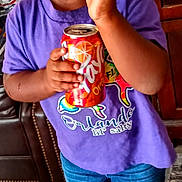 La'Talia Hines is registered to the contest to win money with this photo: child, toddler, smiling, holding, soda_can, orange_soda, purple_shirt, denim_shorts, hair_bows, indoor, living_room, furniture, slippers, happy, cute, person, portrait, floor, wooden_floor, cabinet
