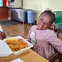 child, girl, birthday, smile, dress, food, fries, chicken_tenders, table, restaurant, lunch, happy, indoor, hairstyle, bow, takeout_container, wooden_table, person, celebration, casual