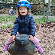 Lily participe au concours pour gagner de l'argent avec cette photo : child, toddler, helmet, jacket, outdoor, playground, wooden_sculpture, duck, smiling, pink_pants, purple_shoes, fence, trees, leaves, bike, happy, nature, daylight, person, play